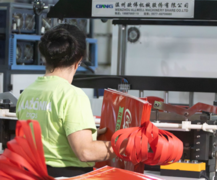 Trabalhadores operam máquina industrial produzindo sacolas vermelhas.