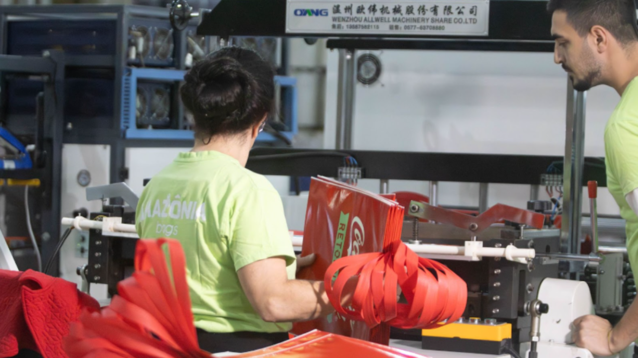 Trabalhadores operam máquina industrial produzindo sacolas vermelhas.