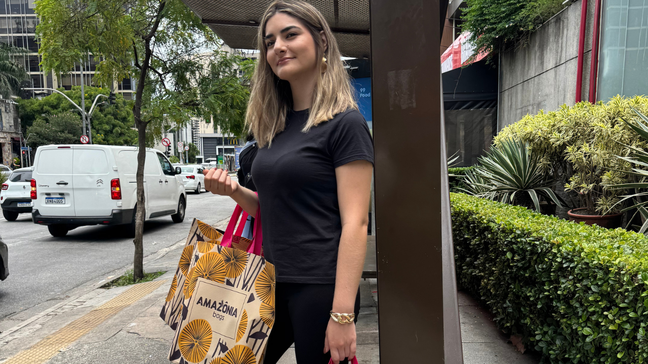 Mulher em área urbana segurando sacolas retornáveis estampadas após realizar compras.