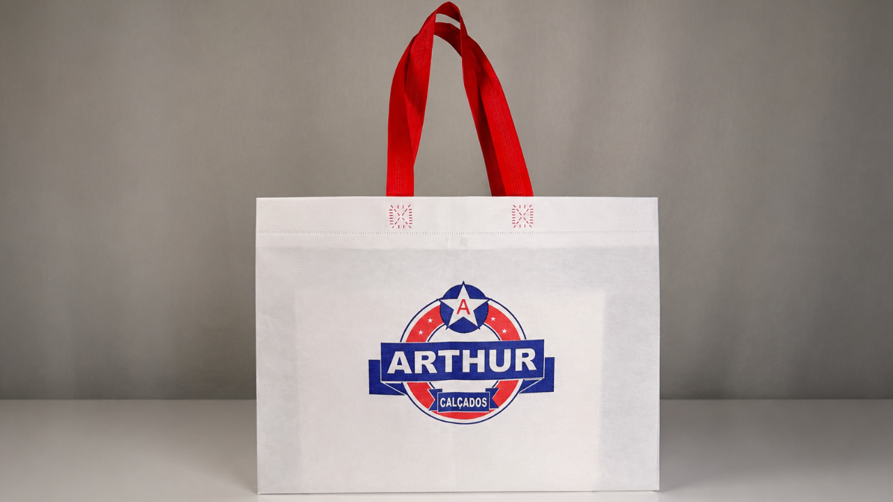 Sacola reutilizável branca com alças vermelhas e logotipo da Arthur Calçados estampado na parte frontal.