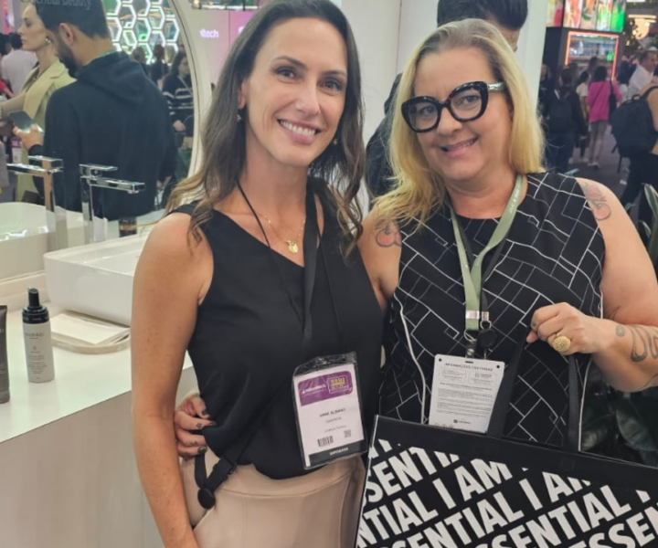 Duas mulheres sorrindo em um evento, ao lado de um estande de produtos de beleza.