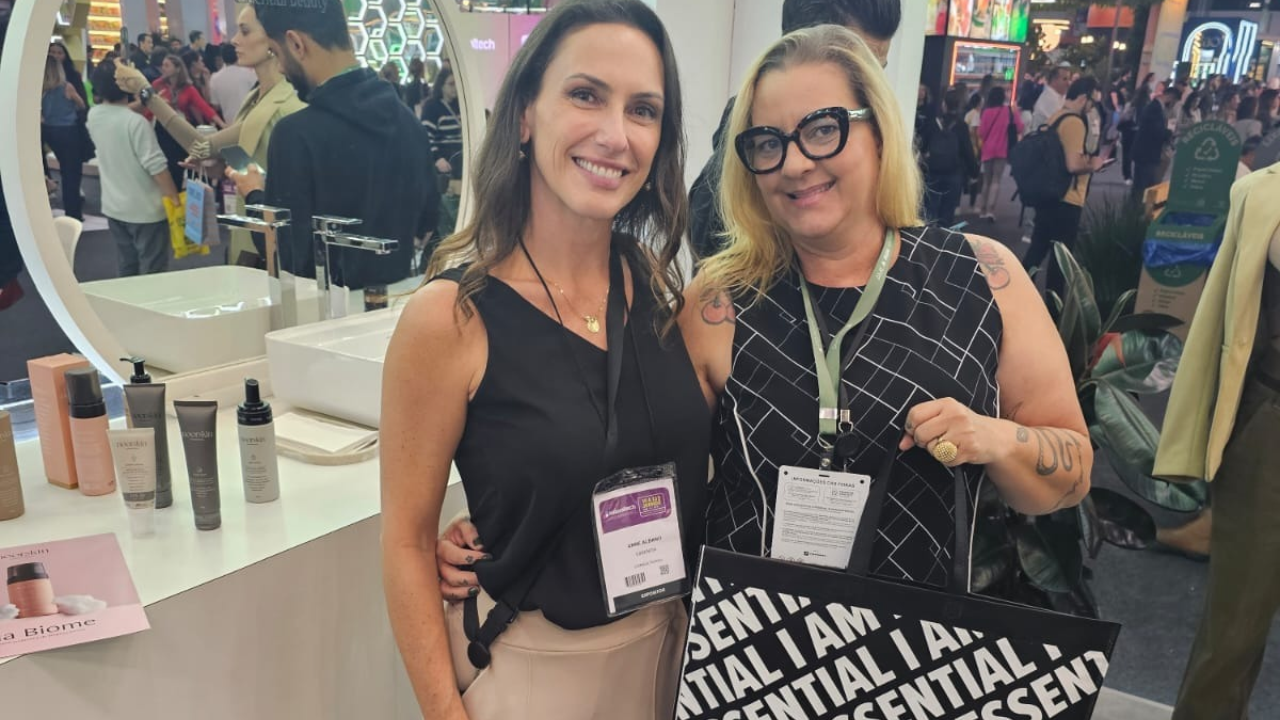 Duas mulheres sorrindo em um evento, ao lado de um estande de produtos de beleza.