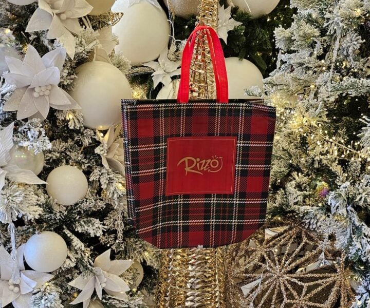 Sacolas personalizadas para evento são ótimas para divulgação orgânica. N imagem, temos uma sacola personalizada com cores natalinas (verde, vermelho e dourado) e logo da marca no centro, posicionada entre decorações de natal brancas.