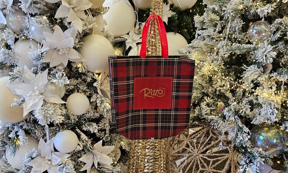 Sacolas personalizadas para evento são ótimas para divulgação orgânica. N imagem, temos uma sacola personalizada com cores natalinas (verde, vermelho e dourado) e logo da marca no centro, posicionada entre decorações de natal brancas.